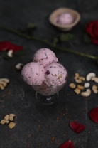 A glass bowl filled with three scoops of pink ice cream, garnished with nuts and rose petals, sits on a dark surface. Scattered cashew nuts and rose petals surround the bowl, with a red rose and another small bowl containing a single scoop of ice cream in the background.