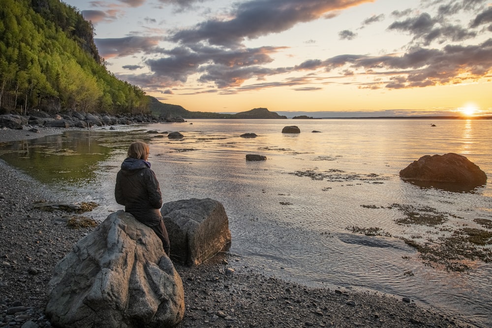 Frau in schwarzer Jacke sitzt tagsüber auf Felsen am Meer