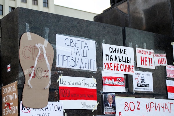 A collection of protest signs and messages taped onto a dark wall. The signs are written in various languages, with some featuring slogans, political messages, and images. The messages appear to express dissent and political activism.