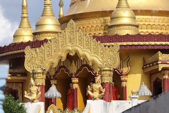 An ornate temple with golden spires and intricate carvings. The architecture features detailed gold embellishments and statues, with red accents. The overall design showcases traditional and religious artistry, with elements of spirituality.