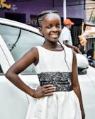 A young girl is smiling, standing in front of a white car. She is wearing a white dress with a black lace belt. Her hair is styled in braids, and she is posing with one hand on her hip. In the background, there is a person in a cap, and a hanging toy chicken is visible.
