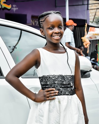 A young girl is smiling, standing in front of a white car. She is wearing a white dress with a black lace belt. Her hair is styled in braids, and she is posing with one hand on her hip. In the background, there is a person in a cap, and a hanging toy chicken is visible.