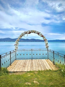 A wooden platform with a decorative metal railing extends over a serene, blue lake. An archway adorned with flowers stands at the end of the platform, with mountains visible in the distance under a partly cloudy sky.