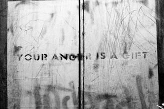 A monochrome image depicts a rough, scratched surface with the words 'YOUR ANGER IS A GIFT' stenciled in bold capital letters. The background appears to be metal with various markings and scratches.