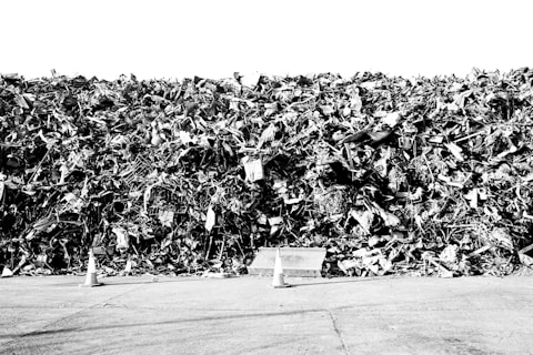 A massive pile of metal and debris, possibly scrap or junk, arranged in a large heap. Several traffic cones are placed in front of the pile, and a largely empty concrete or asphalt surface is visible in the foreground. The image is in black and white, giving it a stark and dramatic feel.