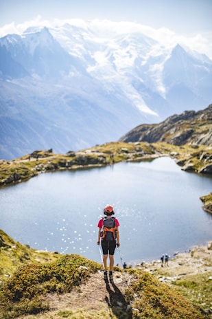 man in red jacket and black pants standing on rock near lake during daytime