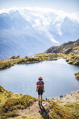 man in red jacket and black pants standing on rock near lake during daytime