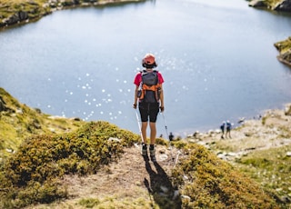 man in red jacket and black pants standing on rock near lake during daytime