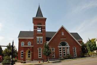 A red brick building with a tall bell tower and pointed roof stands prominently. The structure features multiple arched windows and a main entrance with a small awning. Trees and shrubs are neatly arranged around the building, and the sky is clear.