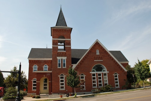 A red brick building with a tall bell tower and pointed roof stands prominently. The structure features multiple arched windows and a main entrance with a small awning. Trees and shrubs are neatly arranged around the building, and the sky is clear.
