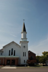 A tall, white church building with a prominent steeple featuring a cross at the top. The structure has arched windows and a blend of brick and white exterior elements. Trees and a street are visible in the foreground under a clear blue sky.