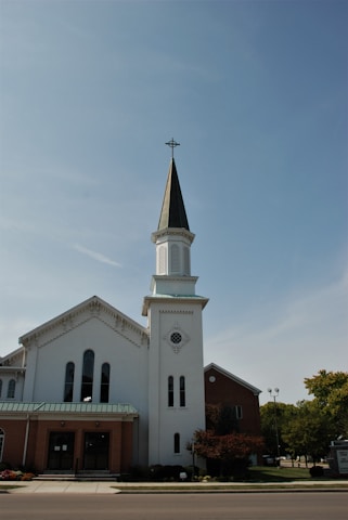 A tall, white church building with a prominent steeple featuring a cross at the top. The structure has arched windows and a blend of brick and white exterior elements. Trees and a street are visible in the foreground under a clear blue sky.