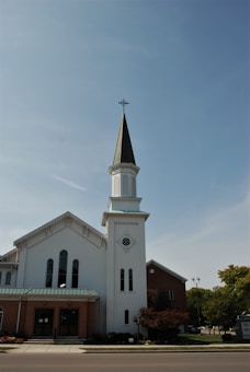 A tall, white church building with a prominent steeple featuring a cross at the top. The structure has arched windows and a blend of brick and white exterior elements. Trees and a street are visible in the foreground under a clear blue sky.