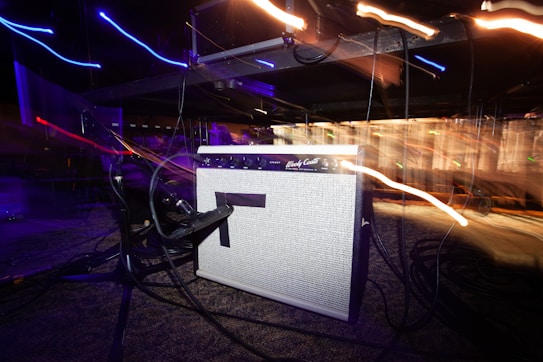 An amplifier is positioned on a carpeted floor with several cables plugged into it. There are light trails in various colors, suggesting movement and energy, against a dark background.