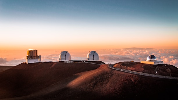 Three observatories with large domes are positioned on a mountain peak above the clouds under a vivid orange and blue sky during sunset. The landscape features a rugged, reddish-brown terrain while a distant island or landmass is visible on the horizon.