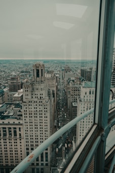 A high-angle view of an urban cityscape featuring tall historic buildings lining a busy street. The scene is viewed through a glass window from a high vantage point, offering a panoramic view of the city. The city extends far into the horizon under a cloudy sky, creating a sense of depth and vastness.