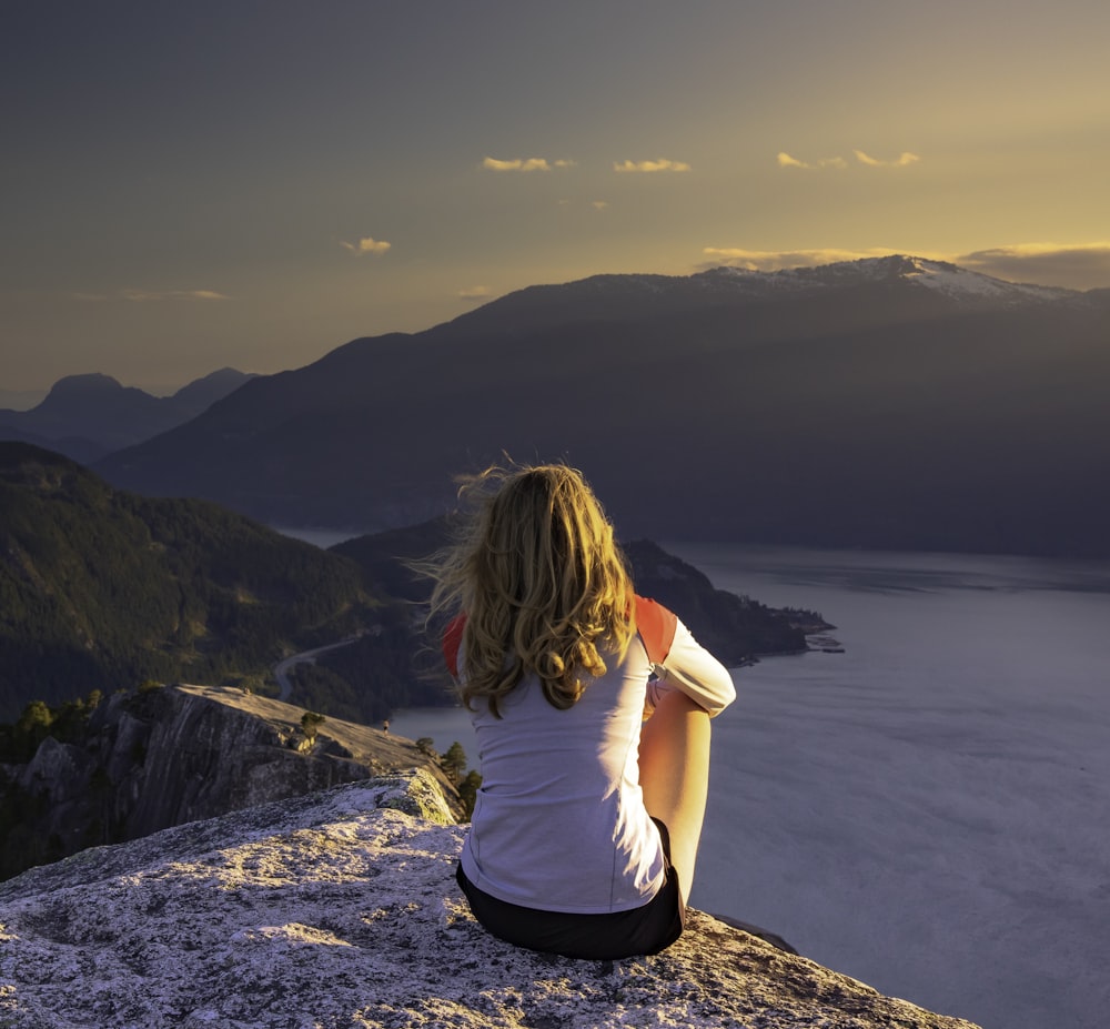 woman in white t-shirt sitting on rock formation near body of water during daytime