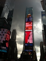 Tall skyscrapers under a cloudy sky surround a vibrant digital billboard displaying various advertisements in a bustling urban setting.