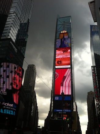 Tall skyscrapers under a cloudy sky surround a vibrant digital billboard displaying various advertisements in a bustling urban setting.