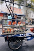 A man stands behind a mobile food cart equipped with glass panels and various containers filled with ingredients. He appears to be preparing or serving food on a city street. The cart has signage with text in a non-English language. In the background, there are buildings, signage, and a parked bicycle.