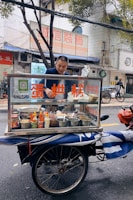 A man stands behind a mobile food cart equipped with glass panels and various containers filled with ingredients. He appears to be preparing or serving food on a city street. The cart has signage with text in a non-English language. In the background, there are buildings, signage, and a parked bicycle.