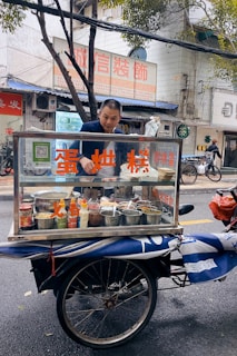 A man stands behind a mobile food cart equipped with glass panels and various containers filled with ingredients. He appears to be preparing or serving food on a city street. The cart has signage with text in a non-English language. In the background, there are buildings, signage, and a parked bicycle.