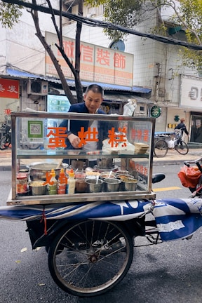 A man stands behind a mobile food cart equipped with glass panels and various containers filled with ingredients. He appears to be preparing or serving food on a city street. The cart has signage with text in a non-English language. In the background, there are buildings, signage, and a parked bicycle.