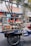 A man stands behind a mobile food cart equipped with glass panels and various containers filled with ingredients. He appears to be preparing or serving food on a city street. The cart has signage with text in a non-English language. In the background, there are buildings, signage, and a parked bicycle.