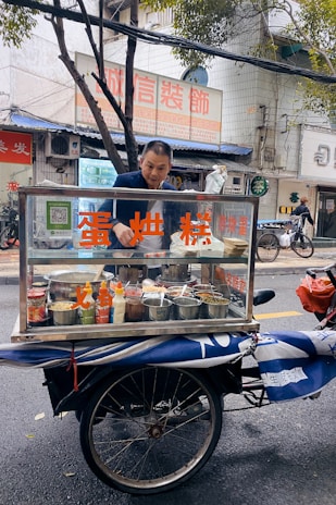 A man stands behind a mobile food cart equipped with glass panels and various containers filled with ingredients. He appears to be preparing or serving food on a city street. The cart has signage with text in a non-English language. In the background, there are buildings, signage, and a parked bicycle.