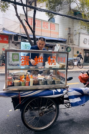 A man stands behind a mobile food cart equipped with glass panels and various containers filled with ingredients. He appears to be preparing or serving food on a city street. The cart has signage with text in a non-English language. In the background, there are buildings, signage, and a parked bicycle.