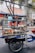 A man stands behind a mobile food cart equipped with glass panels and various containers filled with ingredients. He appears to be preparing or serving food on a city street. The cart has signage with text in a non-English language. In the background, there are buildings, signage, and a parked bicycle.