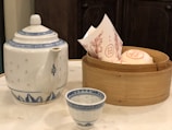 A traditional tea set consisting of a large, ornate teapot with a blue pattern, accompanied by a matching small teacup. Nearby, there is a bamboo steamer basket containing paper-wrapped items possibly representing steamed buns.