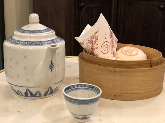 A traditional tea set consisting of a large, ornate teapot with a blue pattern, accompanied by a matching small teacup. Nearby, there is a bamboo steamer basket containing paper-wrapped items possibly representing steamed buns.