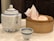 A traditional tea set consisting of a large, ornate teapot with a blue pattern, accompanied by a matching small teacup. Nearby, there is a bamboo steamer basket containing paper-wrapped items possibly representing steamed buns.