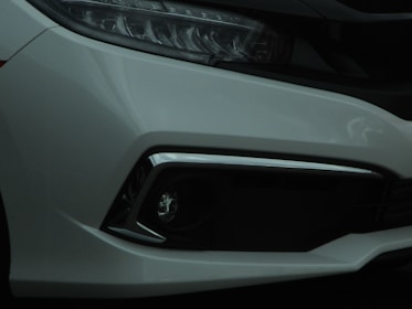 Close-up of a car's front bumper and headlight area. The design features sleek lines with a modern aesthetic, showing part of the grille and the headlight assembly.