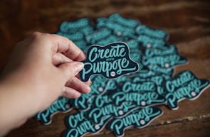 A hand is holding a sticker with the words 'Create with Purpose' written in a stylish script. Multiple similar stickers are scattered on a wooden surface in the background.