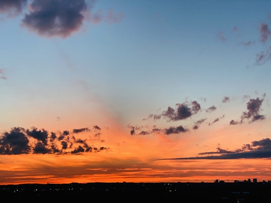 The image captures a vibrant sunset with a mix of orange and pink hues across the horizon. Scattered clouds add texture against the transitioning sky, which fades from deep orange to a soft blue. Silhouetted structures appear at the bottom, providing contrast to the colorful sky.