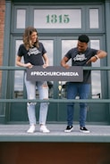 Two people stand on a balcony holding a sign with the text '#PROCHURCHMEDIA', both wearing matching 'Create Purpose' t-shirts. The background shows a building door with the number '1315'.