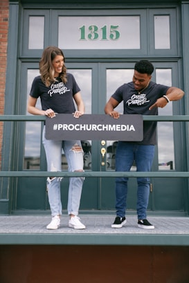 Two people stand on a balcony holding a sign with the text '#PROCHURCHMEDIA', both wearing matching 'Create Purpose' t-shirts. The background shows a building door with the number '1315'.
