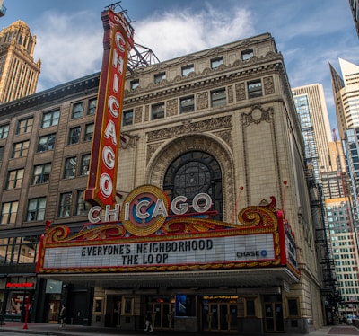 A historic theater marquee with the iconic CHICAGO sign in bold red letters is prominently displayed. The building has ornate architectural details with an arched window above the entrance. The marquee displays the text 'EVERYONE'S NEIGHBORHOOD THE LOOP', and surrounding skyscrapers are visible in the background.