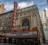 A historic theater marquee with the iconic CHICAGO sign in bold red letters is prominently displayed. The building has ornate architectural details with an arched window above the entrance. The marquee displays the text 'EVERYONE'S NEIGHBORHOOD THE LOOP', and surrounding skyscrapers are visible in the background.