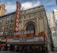 A historic theater marquee with the iconic CHICAGO sign in bold red letters is prominently displayed. The building has ornate architectural details with an arched window above the entrance. The marquee displays the text 'EVERYONE'S NEIGHBORHOOD THE LOOP', and surrounding skyscrapers are visible in the background.