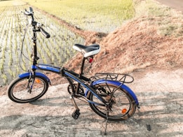 A blue folding bicycle is parked on a concrete path beside a rice field. The bike is positioned with its side stand down, and the handlebars are turned slightly. The rice field in the background is lush and green, with neat rows of young plants in water. The surrounding area is grassy and sunlit, casting clear shadows of the bicycle on the ground.