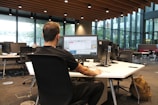 A person is seated at a large desk in an open office space, working on a computer. The office features modern furniture and has large windows that allow natural light to fill the room. Several computer monitors are set up, and there is a backpack resting on the floor next to the desk.