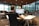 A person is seated at a large desk in an open office space, working on a computer. The office features modern furniture and has large windows that allow natural light to fill the room. Several computer monitors are set up, and there is a backpack resting on the floor next to the desk.