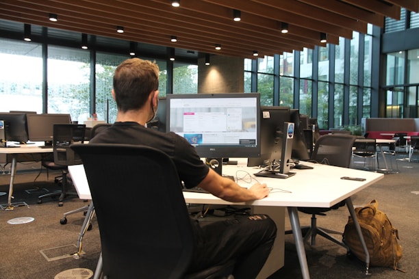 A person is seated at a large desk in an open office space, working on a computer. The office features modern furniture and has large windows that allow natural light to fill the room. Several computer monitors are set up, and there is a backpack resting on the floor next to the desk.