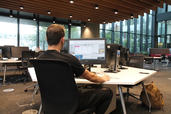 A person is seated at a large desk in an open office space, working on a computer. The office features modern furniture and has large windows that allow natural light to fill the room. Several computer monitors are set up, and there is a backpack resting on the floor next to the desk.