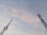 Two telecommunications towers extend into the sky, silhouetted against a backdrop of soft, pastel-colored clouds. The towers display intricate lattice structures with panels and antennas mounted at various levels.