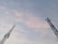 Two telecommunications towers extend into the sky, silhouetted against a backdrop of soft, pastel-colored clouds. The towers display intricate lattice structures with panels and antennas mounted at various levels.