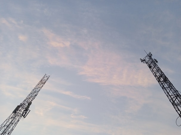 Two telecommunications towers extend into the sky, silhouetted against a backdrop of soft, pastel-colored clouds. The towers display intricate lattice structures with panels and antennas mounted at various levels.